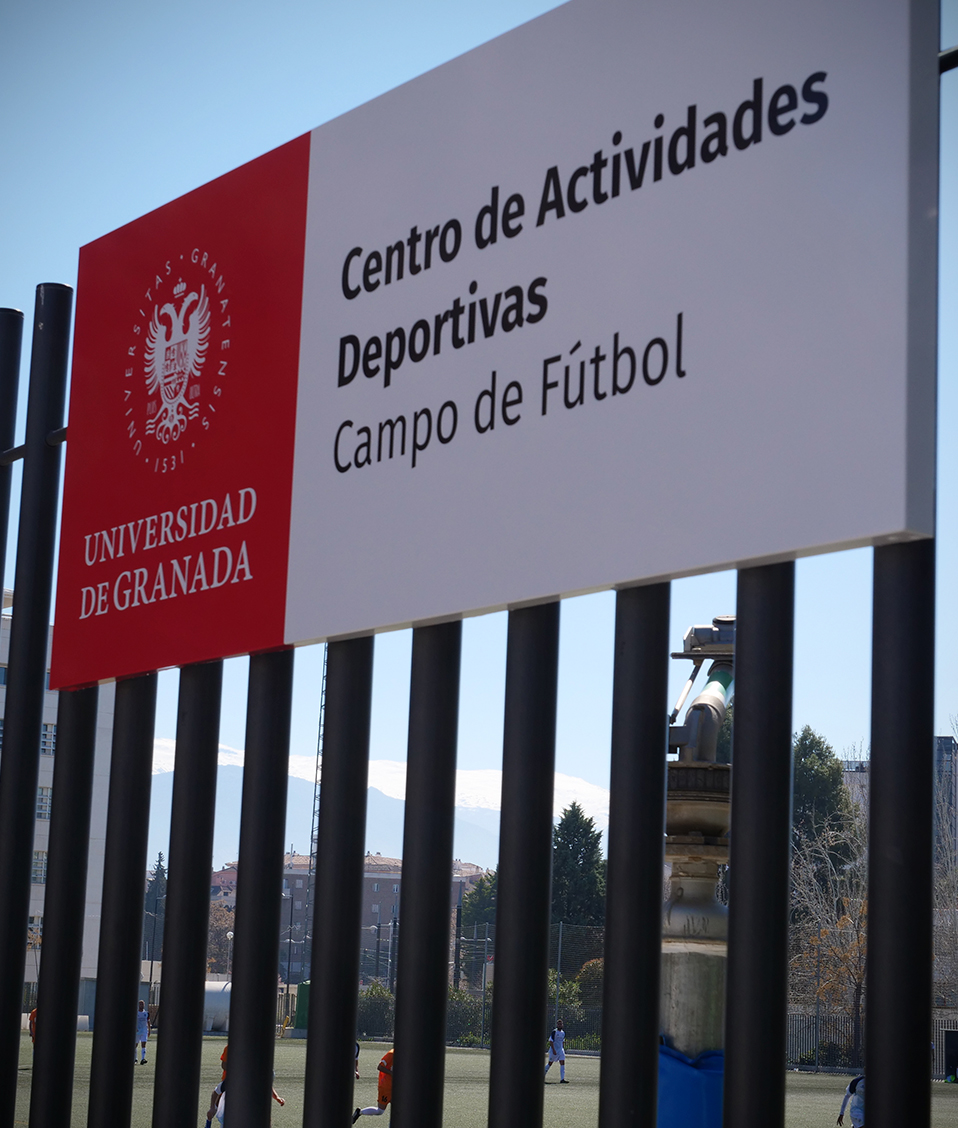 Cartel del campo de fútbol del Centro de Actividades Deportivas del Campus de Fuentenueva colocado sobre una valla tras la que se ven los jugadores y algunos edificios